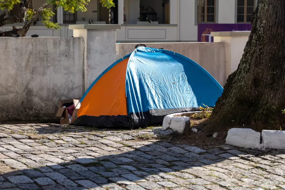 A orange-and-blue tent set up next to a tree in a paved courtyard and a cardboard box of posessions next to the tent entrance.